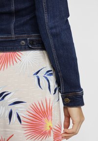 Chaqueta de mezclilla azul oscuro con un puño que presenta un botón con logo, combinada con una falda de color claro con estampado floral.
