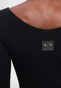 Top nero a costine con scollatura ampia. Presenta un'etichetta rettangolare con il logo "A/X" e il testo "BLACK EDITION". Tessuto liscio, design aderente.