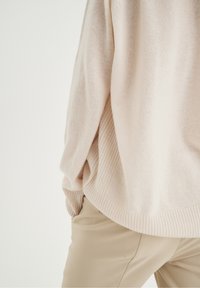 Pull ample beige clair avec ourlet et manches côtelés porté sur un pantalon beige assorti, mains dans les poches, fond neutre.