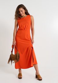 Vestido maxi sin mangas naranja con abertura frontal, tejido suave y un corsé ajustado. Combinado con sandalias de tacón marrones y un bolso con flecos.