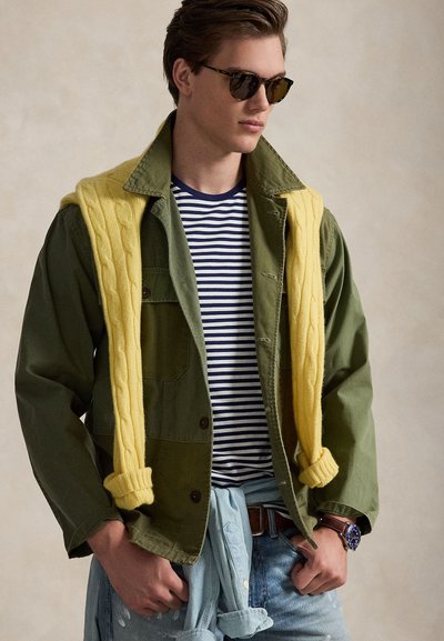 Νεαρός άνδρας που φοράει γυαλιά ηλίου με σχέδιο χελώνας, ελαιμοβρύχη πράσινη ζακέτα, μπλε ριγέ πουκάμισο, κίτρινο πουλόβερ με πλεξούδες ριγμένο στους ώμους και ρολόι.