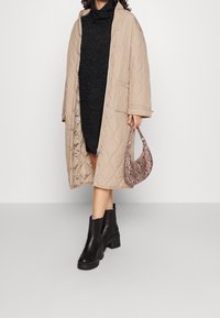 Beige kviltad lång kappa med tryckknappar, bärs över en mörk tröjdress. Svarta ankelstövlar med tjocka sulor och en rosa orm-lädersväska.