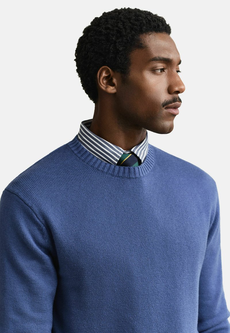 Mann mit kurzen lockigen Haaren, der einen blauen Pullover über einem blau-weiß gestreiften Hemd und einer grün gestreiften Krawatte trägt und nach rechts schaut.
