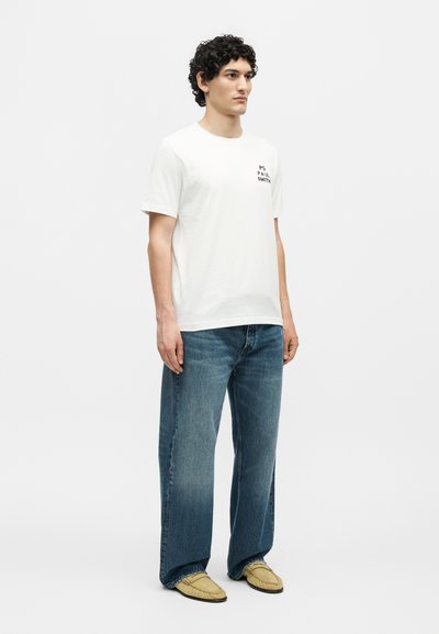 Άνδρας με σγουρά μαλλιά που φοράει λευκό T-shirt PS Paul Smith, φαρδιά μπλε τζιν και μπεζ μοκασίνια, στέκεται μπροστά σε ανοιχτόχρωμο φόντο.