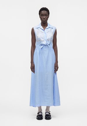Femme portant une robe maxi bleu clair sans manches avec des découpes florales brodées, taille nouée, chaussettes blanches et chaussures noires à boucle, debout.