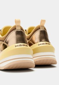 Zapatillas de color bronce metálico con detalles en amarillo, presentan una parte trasera texturizada, suela con rayas blancas y beige, y una lengüeta con la marca.