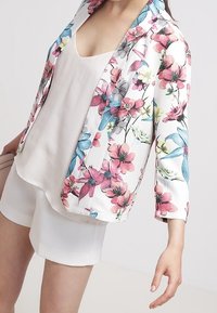 Kwiecista marynarka z białym tłem, różowymi i niebieskimi kwiatami, rękawami o długości trzy czwarte oraz luźnym krojem, zestawiona z jasnym topem.