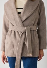 Cappotto avvolgente in misto lana beige con ampi risvolti, una cintura e una finitura testurizzata. Presenta tasche laterali ed è abbinato a un maglione a righe sotto.