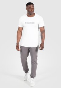 Valkoinen lyhythihainen t-paita, jossa on "SMILODOX"-logo, yhdistettynä harmaisiin verkkareihin ja valkoisiin lenkkareihin, jossa on slim fit ja pehmeä tekstuuri.