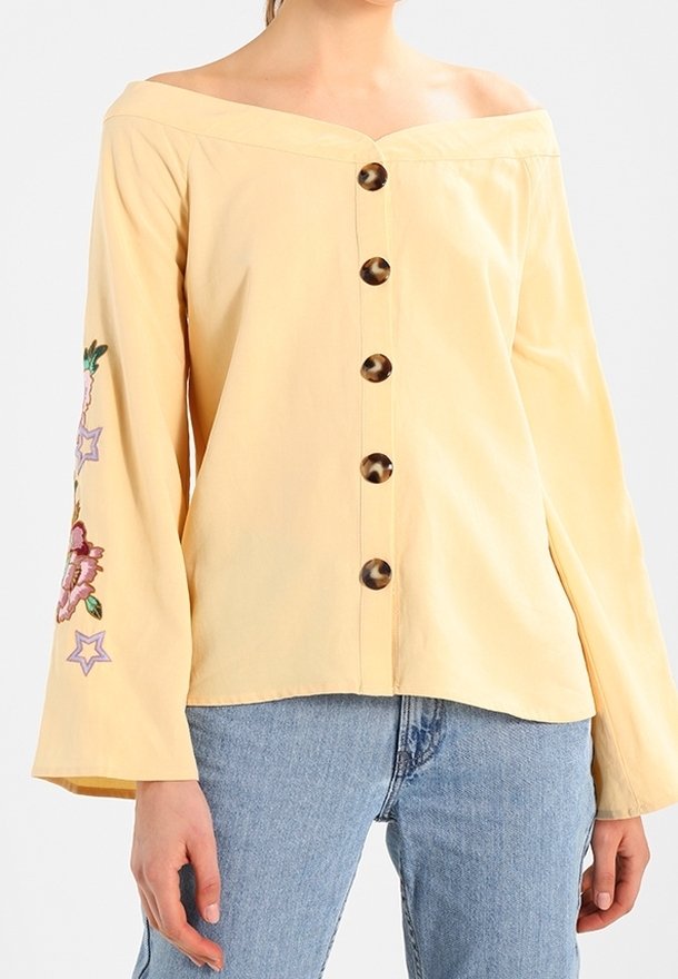 Femme portant un chemisier jaune pâle à épaules dénudées avec de gros boutons et une broderie florale sur une manche, associé à un jean bleu clair.
