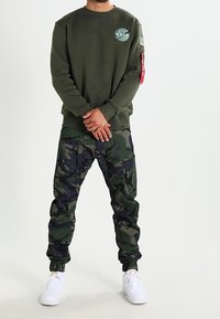 Felpa verde con logo NASA, abbinata a pantaloni cargo a fantasia mimetica in verde e blu navy. Scarpe da ginnastica bianche completano l'outfit.