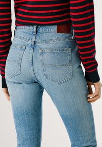 Heledad sinine teksapüksid kõrge vöökohaga, kaks tagataskut ja nahast brändi silt. Kombineeritud musta ja punase triibulise svetriga.