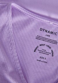 Lavendel ribbad tyg med V-ringning, med tryckta etiketter som säger "DYNAMIC", "SIZE S" och fetstilta beskrivande termer.