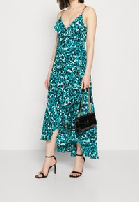 Robe maxi en teal avec un imprimé dispersé, présentant des détails en volants, des bretelles fines et une fente haute. Associée à des talons noirs à lanières et un sac à main.