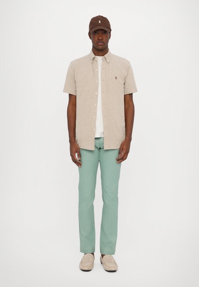 Camisa de botones corta de manga corta color beige con una camiseta gris clara debajo, combinada con pantalones verde menta y mocasines beige. Gorro marrón con logotipo.