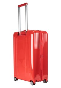 Maleta dura roja y brillante con surcos verticales, asa extensible plateada y cuatro ruedas negras para un desplazamiento suave.
