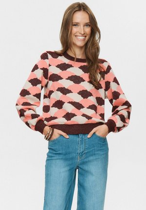 Eine Frau lächelt, mit den Händen in den Taschen, trägt einen weit geschnittenen Pullover mit pinken, braunen und beigen Mustern sowie blaue Jeans vor einem weißen Hintergrund.