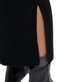 Czarna dzianinowa sukienka z bocznym rozcięciem, odsłaniająca jasnoszary prążkowany skarpetę i czarny skórzany but o gładkiej fakturze.