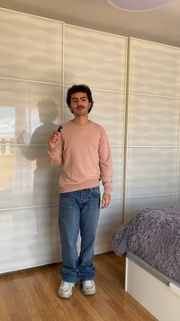 Mladi moški v rožnatem puloverju in kavbojkah drži majhen črn predmet, stoji v prostoru s svetlo obarvano obločno steno in lesenimi tlemi.