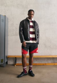 Μαύρο δερμάτινο μπουφάν, ριγέ μπορντώ και λευκό πόλο, μαύρες βερμούδες με κόκκινες λεπτομέρειες, κόκκινες και λευκές ριγέ κάλτσες, μαύρα αθλητικά παπούτσια.