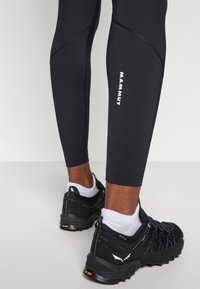 Čierne športové legíny s úzky fit a bielym logom "MAMMUT" na boku, kombinované s čiernymi bežeckými topánkami na trail a bielymi ponožkami.