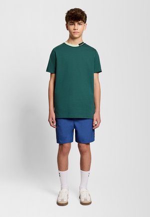 Teenagedreng stående iført en mørkegrøn t-shirt, blå shorts, hvide crew-sokker og beige sneakers foran en ensfarvet lys baggrund.