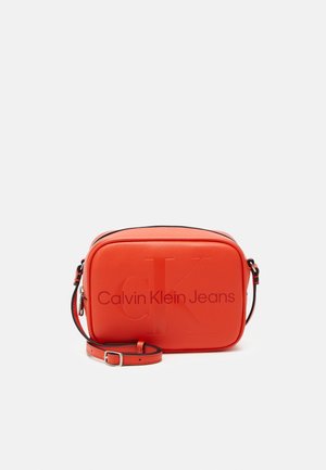 Červená crossbody taška obdélníkového tvaru, hladké textury, s embosovaným logem "CK" a nastavitelným černým popruhem. Zapínání na zip nahoře.