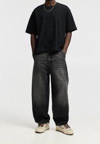 T-shirt noir à manches courtes associé à un jean noir délavé et ample, avec des baskets beiges ornées d'accents marron et de lacets.