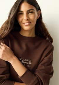 Brauner Pullover aus weichem Material, mit weißer, bestickter Schrift. Rundhalsausschnitt und lockere Passform mit gerippten Bündchen.