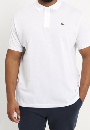 Mann in einem einfachen weißen Poloshirt mit kurzen Ärmeln und grünem Krokodil-Logo auf der Brust, trägt dunkle Hosen und steht mit einer Hand in der Tasche.