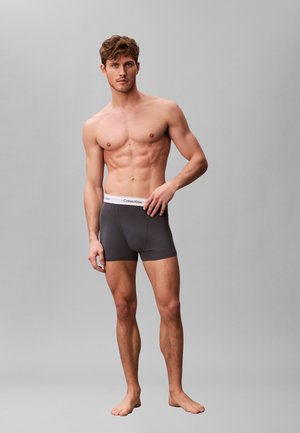 Shirtloser Mann, der dunkelgraue Calvin Klein Boxershorts trägt, steht barfuß vor einem schlichten grauen Hintergrund.