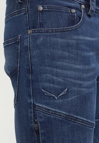 Mörkblå denimjeans med smal passform, synlig sömnad, två framfickor och en diskret logodetalj vid blixtlåsfickan.
