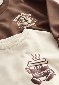 To sweatshirts: en brun med "BONJOUR TODAY" og en tegneserie, en cremefarvet med et "BABYCCINO"-motiv. Begge har teksturerede tryk og ribkantede kraver.