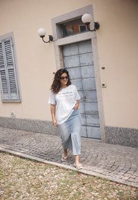 T-shirt graphique blanc avec le texte "besoin de vitamine mer", associé à une longue jupe en denim bleu clair et des tongs blanches. Cheveux bruns, lunettes de soleil.