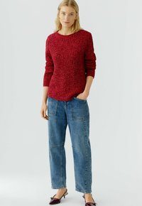 Roter strukturiertes Strickpullover mit Dreiviertelärmeln, kombiniert mit lockeren blauen Jeans mit sichtbaren Nähten und Taschen sowie dunkelroten Schuhen.