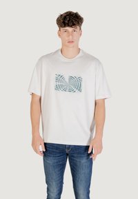 T-shirt bianco in cotone a maniche corte, con una grafica a spirale in testo turchese sul davanti, abbinato a jeans in denim blu.
