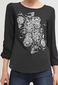 Černý top s dlouhým rukávem a bílou květinovou výšivkou umístěnou na hrudi, nošený s tmavě modrými džíny.