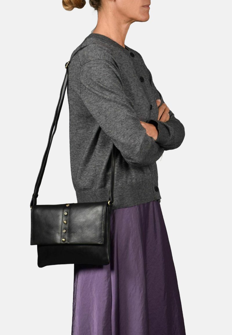 Zwarte leren crossbody tas met een plat ontwerp, heeft drie decoratieve metalen studs en een verstelbare band. Draagt over een grijs trui en een paarse rok.