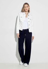 Giacca in denim bianco con tasche frontali, abbinata a pantaloni blu scuro a vestibilità ampia e sneakers bianche. L'outfit presenta un design semplice e un comfort informale.