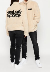 Beżowa polarowa kurtka z wysokim kołnierzem, z czarnym haftowanym napisem "multiply apparel" na plecach oraz małym logo z przodu.