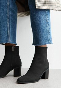 Botas de ante negras hasta el tobillo con punta afilada y tacón cuadrado. Cierre de cremallera lateral, combinadas con jeans vaqueros azul cortos con dobladillos deshilachados.