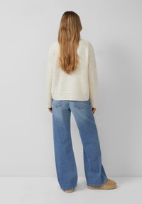 Frau mit langen Haaren, die einen cremefarbenen Strickpullover, eine weit geschnittene blaue Jeans und beige Slipper trägt, steht vor einem schlichten grauen Hintergrund.