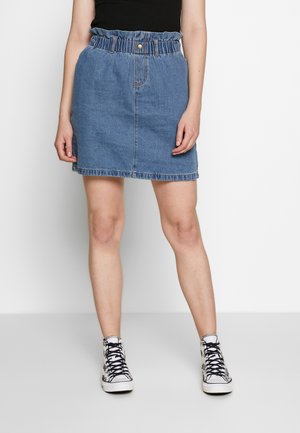 Femme portant une jupe en jean bleue à taille paperbag et des baskets noires et blanches à motifs, debout devant un fond blanc.