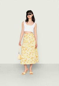 Saia midi floral em amarelo e branco, com um cós ajustado; combinada com um top cropped branco e acessórios amarelos, incluindo sandálias e uma mala.