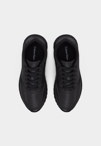 Ζευγάρι μαύρα αθλητικά παπούτσια Calvin Klein με κορδόνια, εμφανιζόμενα από πάνω, με ορατό το όνομα της μάρκας εσωτερικά και στο πλάι.