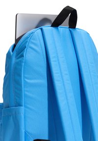 Modrý batoh s nastavitelnými černými popruhy, texturovanou tkaninou a boční kapsou, ze které je částečně vidět stříbrný notebook.