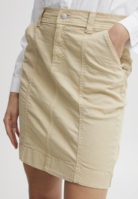 Jupe beige en coton, avec un design ajusté, des coutures visibles, deux poches latérales et une fermeture à bouton à la taille.