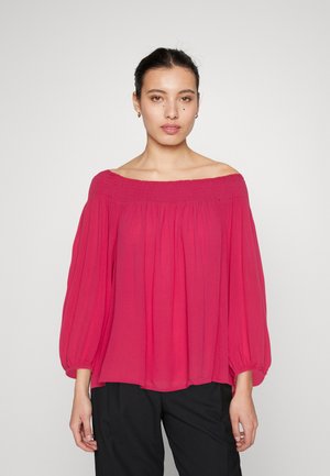 Ροζ μπλούζα με ανοιχτούς ώμους και σμυριδωτή λεπτομέρεια, με μακριά φουσκωτά μανίκια και ελαφρύ ύφασμα, φορεμένη πάνω από μαύρο παντελόνι.