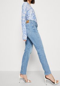 Ljust blå långärmad topp med ett vågigt mönster, kombinerad med ljusblå denimjeans och vita klacksandaler. Noterbara detaljer inkluderar en etikett på jeansen.