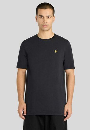 Jonge man staand, vooruit kijkend, draagt een zwart T-shirt met korte mouwen met een klein geel vogel-logo op de borst.